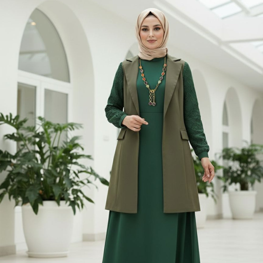 Woman in a green dress with a matching vest standing in a bright indoor setting with plants.