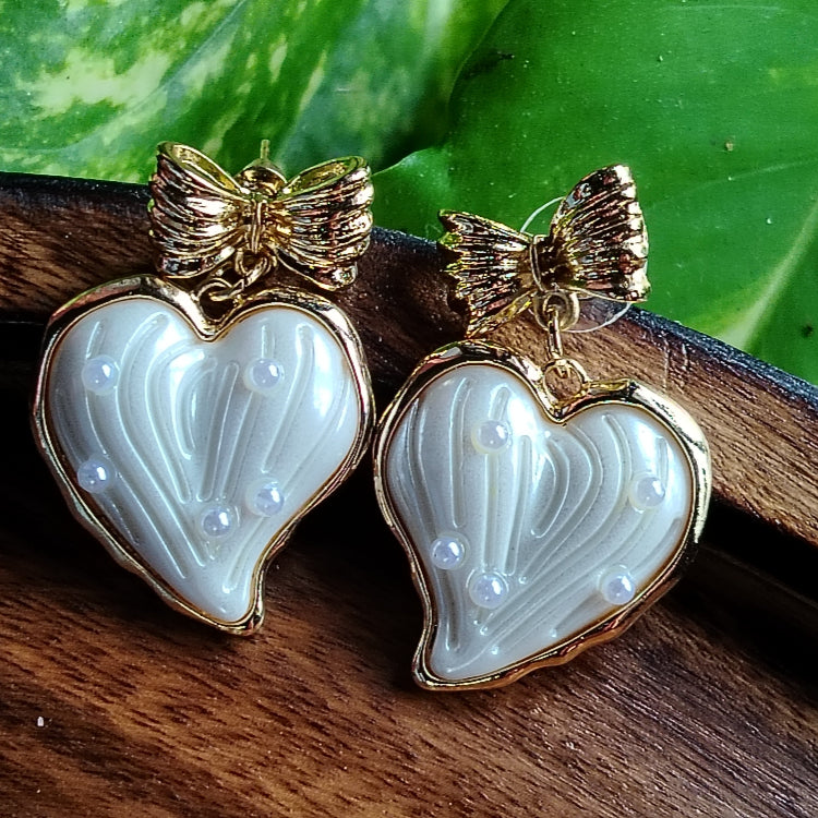 Heart-shaped earrings with Golden Bow Tops on a wooden surface with green leaves in the background