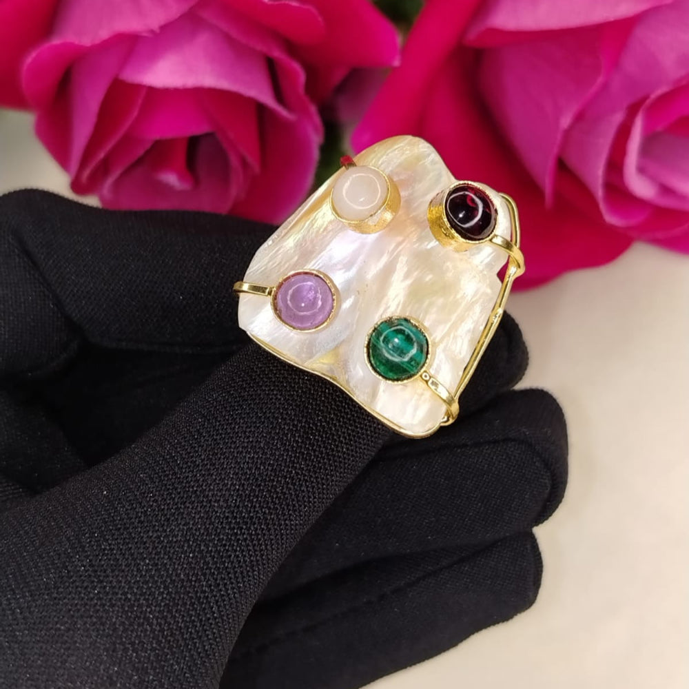 Golden Ring with Pearl Base and colorful Gemstones Held by a Black-gloved hand against Pink Flowers.
