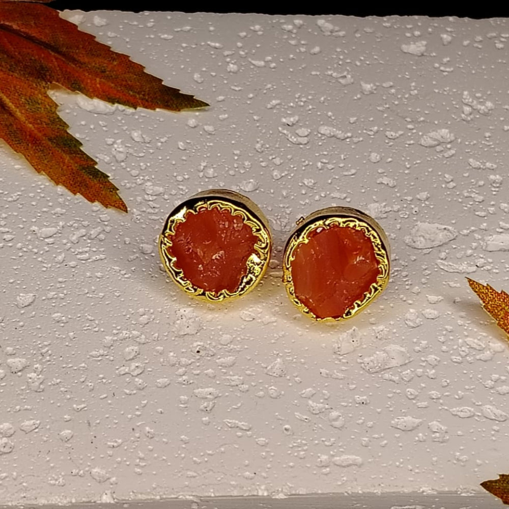 Pair of gold-framed red stone earrings on a textured white surface with autumn leaves.