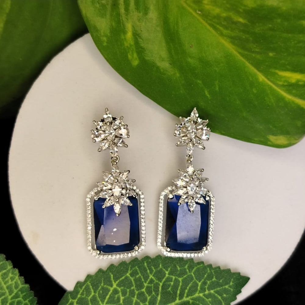 Pair of Silver Polished Earrings with blue gemstones and American Diamonds on a white surface, green leaves in the background.