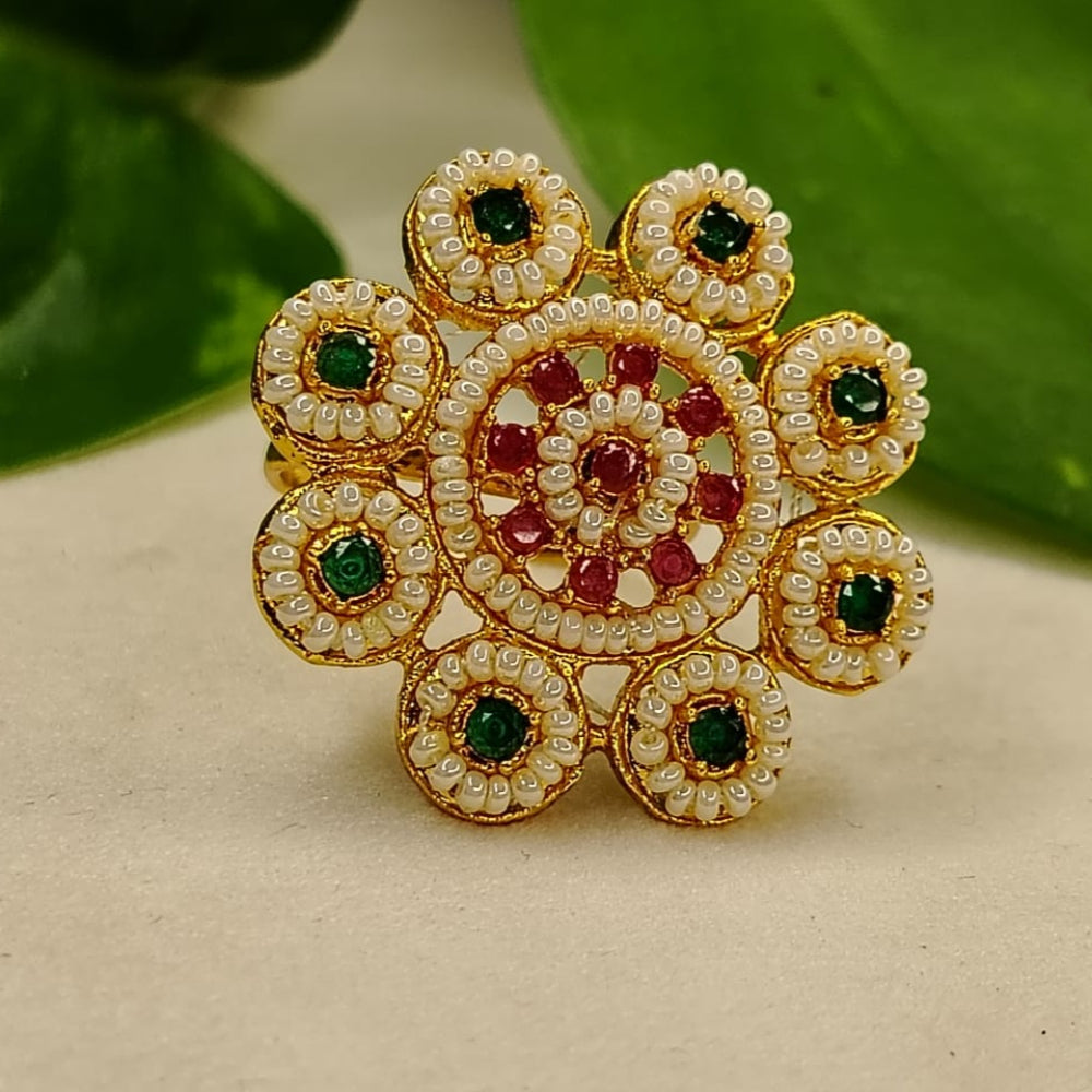 Decorative ring with gold, green, and red stones on a beige surface with green leaves in the background