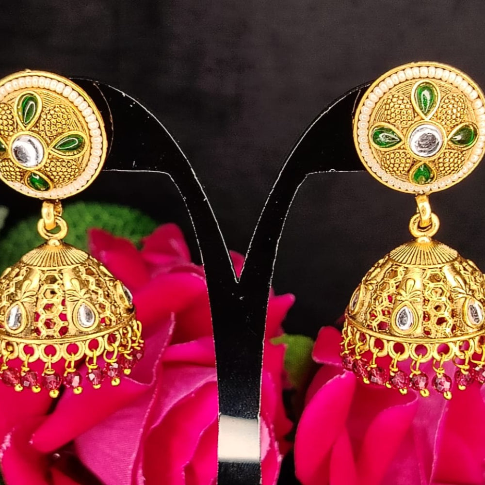 Gold earrings with green and red accents on a black stand against a dark background with pink flowers.