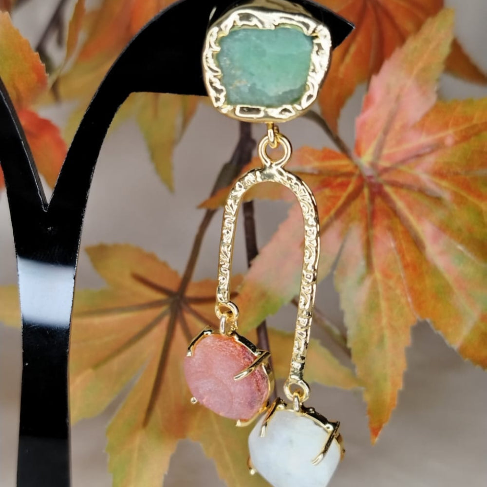 Golden earrings with green, pink, and white gemstones on a stand with autumn leaves in the background