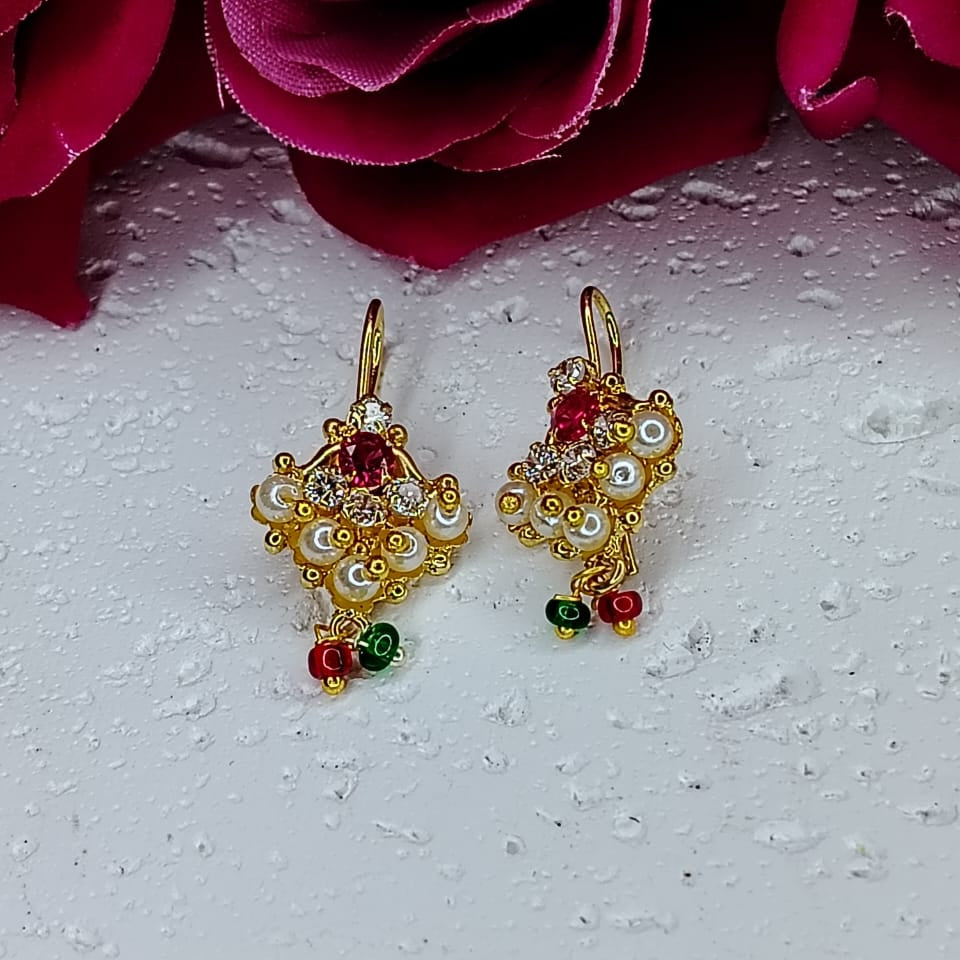 Gold earrings with red and green stones on a white surface with red flowers in the background
