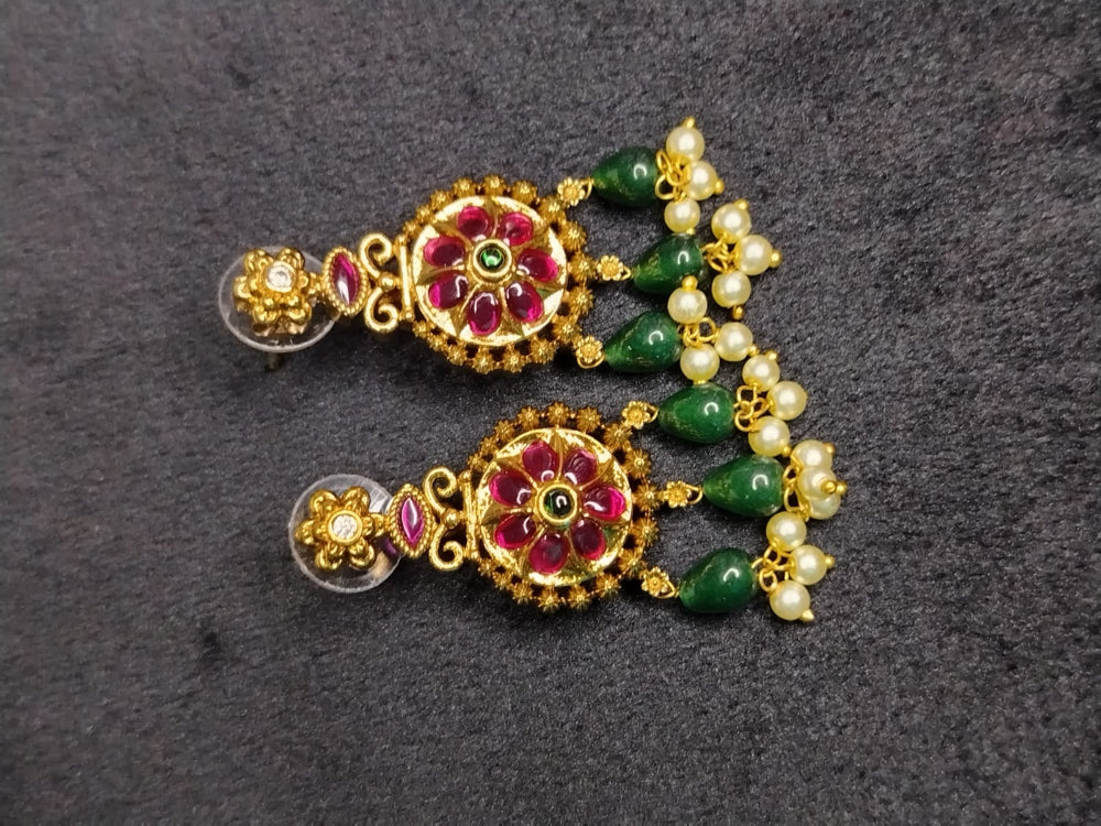 Pair of gold earrings with red gemstones, green beads, and pearl-like elements on a dark gray background.