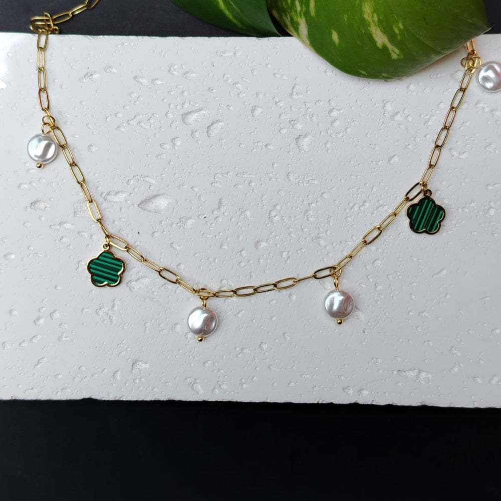 Golden chain necklace with green clover charms and pearls on a white surface with a leafy plant.