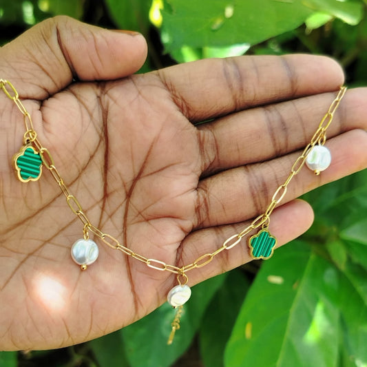 Golden chain necklace with green pendants and pearls held in a hand against a green leafy background