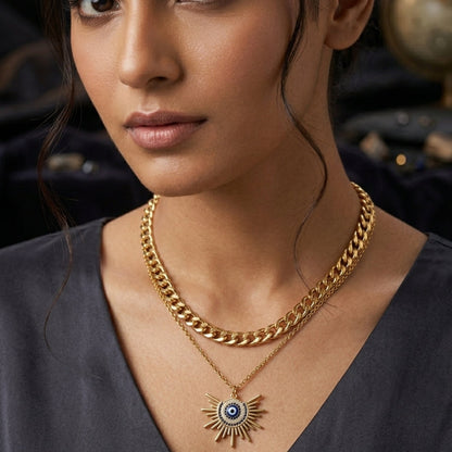 A close-up of a woman wearing a layered gold necklace set, featuring a thick curb chain and a delicate chain with a sunburst pendant containing a blue and white evil eye design.
