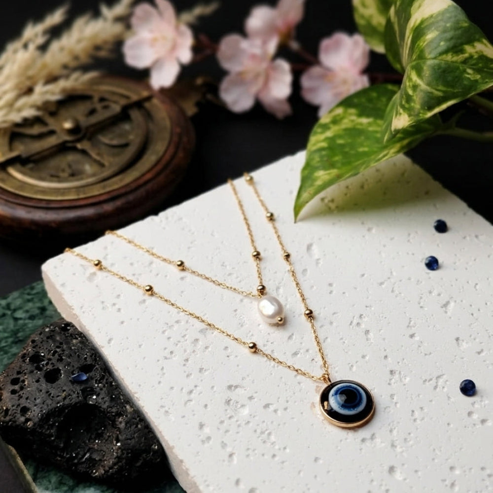 Double-layered golden necklaces displayed on a white, textured stone slab. One layer has a small white pearl, and the other has a circular blue and black evil eye pendant.