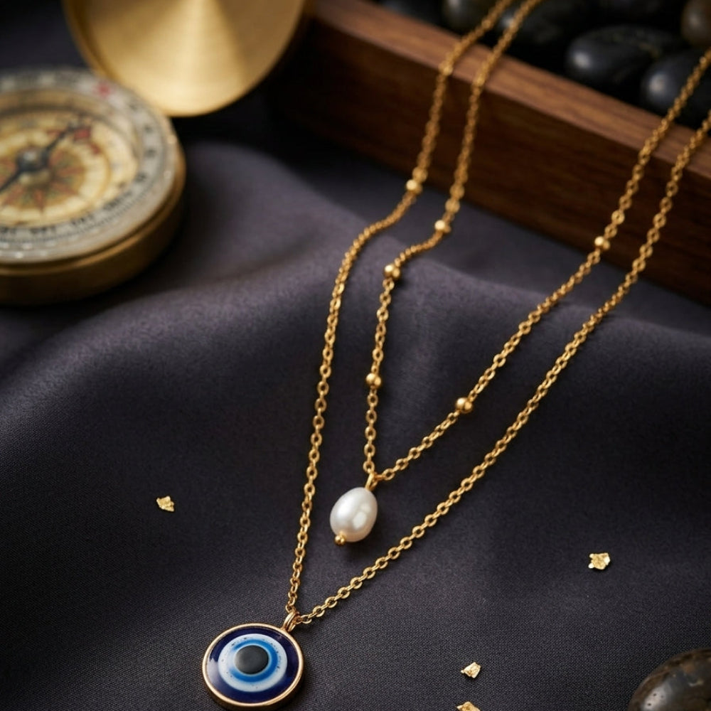 A gold layered necklace set displayed on dark satin fabric. The set includes a delicate gold chain with a white pearl pendant and a longer gold chain featuring a circular evil eye pendant with blue, black, and white concentric details and a gold border.
