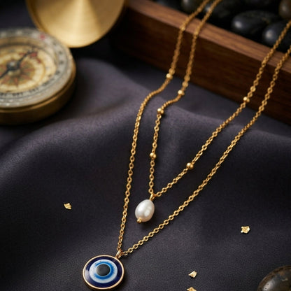 A gold layered necklace set displayed on dark satin fabric. The set includes a delicate gold chain with a white pearl pendant and a longer gold chain featuring a circular evil eye pendant with blue, black, and white concentric details and a gold border.