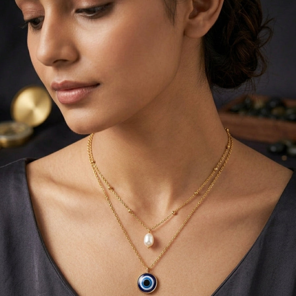 A close-up lifestyle shot of a woman wearing a double-layered gold necklace. The top layer features a small white pearl, and the bottom layer features a circular blue and black evil eye pendant.