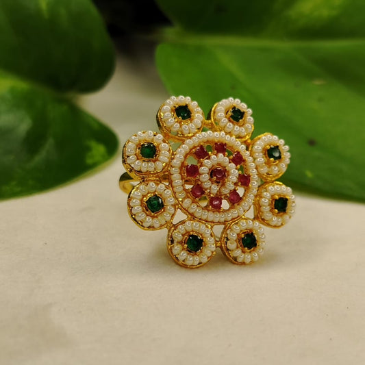 Decorative ring with gold, green, and red stones on a beige surface with green leaves in the background