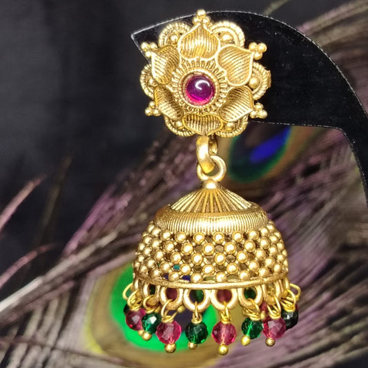 Gold jhumka earring with gemstones on a black stand against a peacock feather background