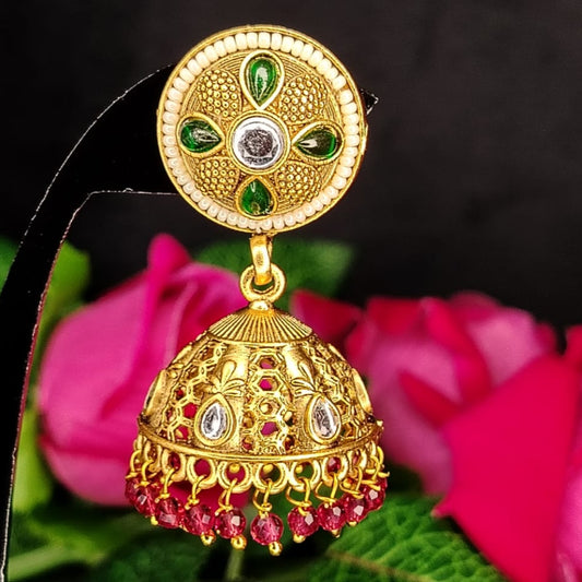 Golden jhumka earring with gemstones on a black stand with pink flowers in the background