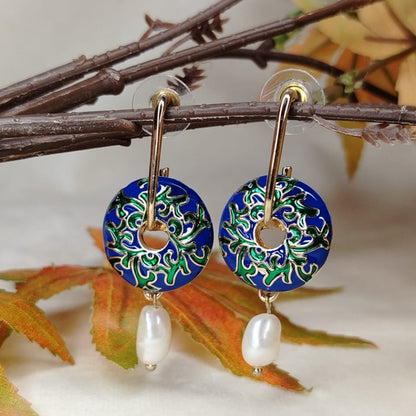 Decorative earrings with blue floral design and pearl beads on a branch with leaves.