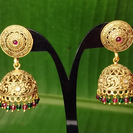 Gold Jhumkas with red and green stones on a black stand against a green leafy background