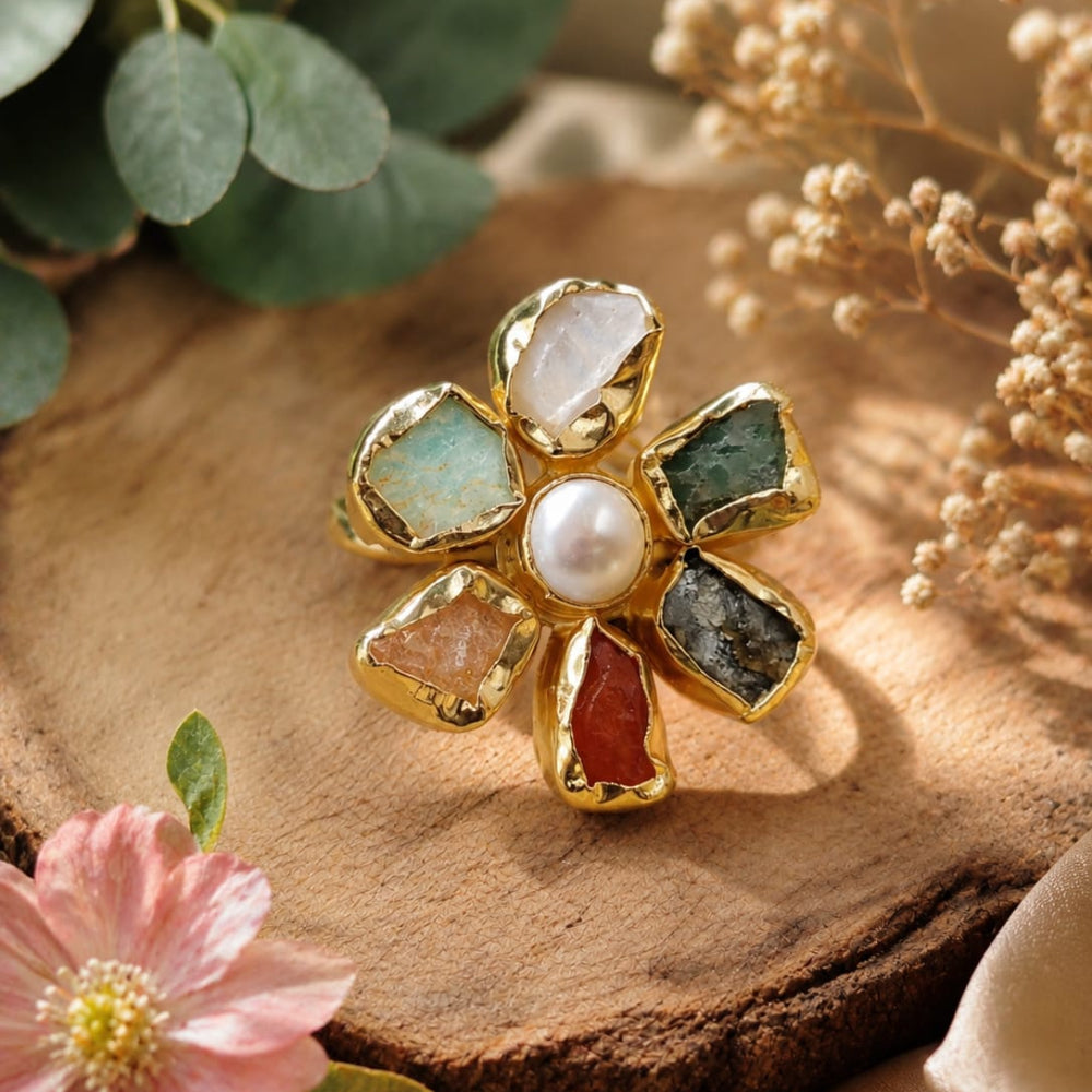 Gold flower-shaped ring with colorful gemstones on a wooden surface with flowers and leaves.