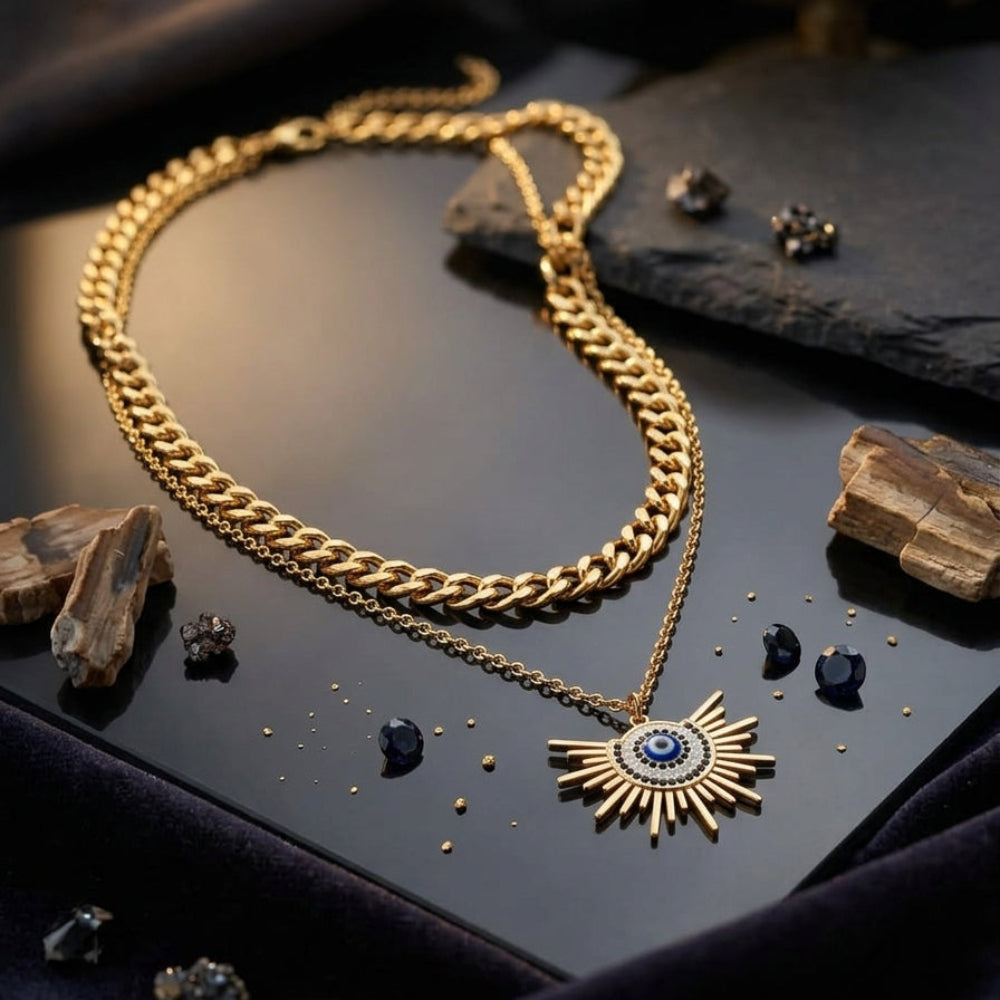A gold-toned double-strand necklace featuring a thick curb chain and a delicate chain with a sunburst pendant containing a blue evil eye motif, displayed on a dark reflective surface with decorative stones and fabric.
